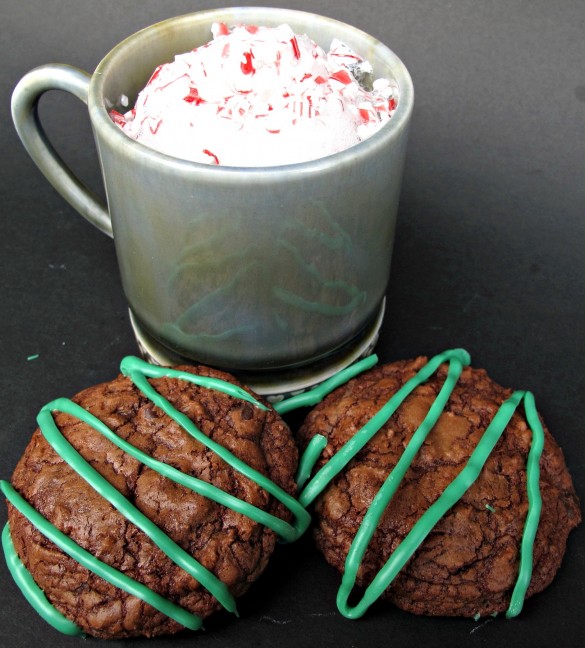 Two mint brownie cookies decorated with green zigzags  with a mug of  cocoa with peppermint bits.