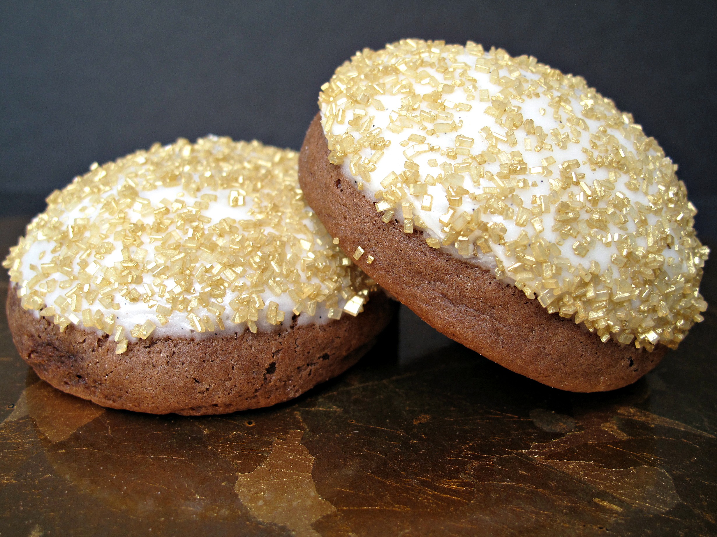 Closeup of two puffy chocolate cookies topped with white icing and gold sugar.