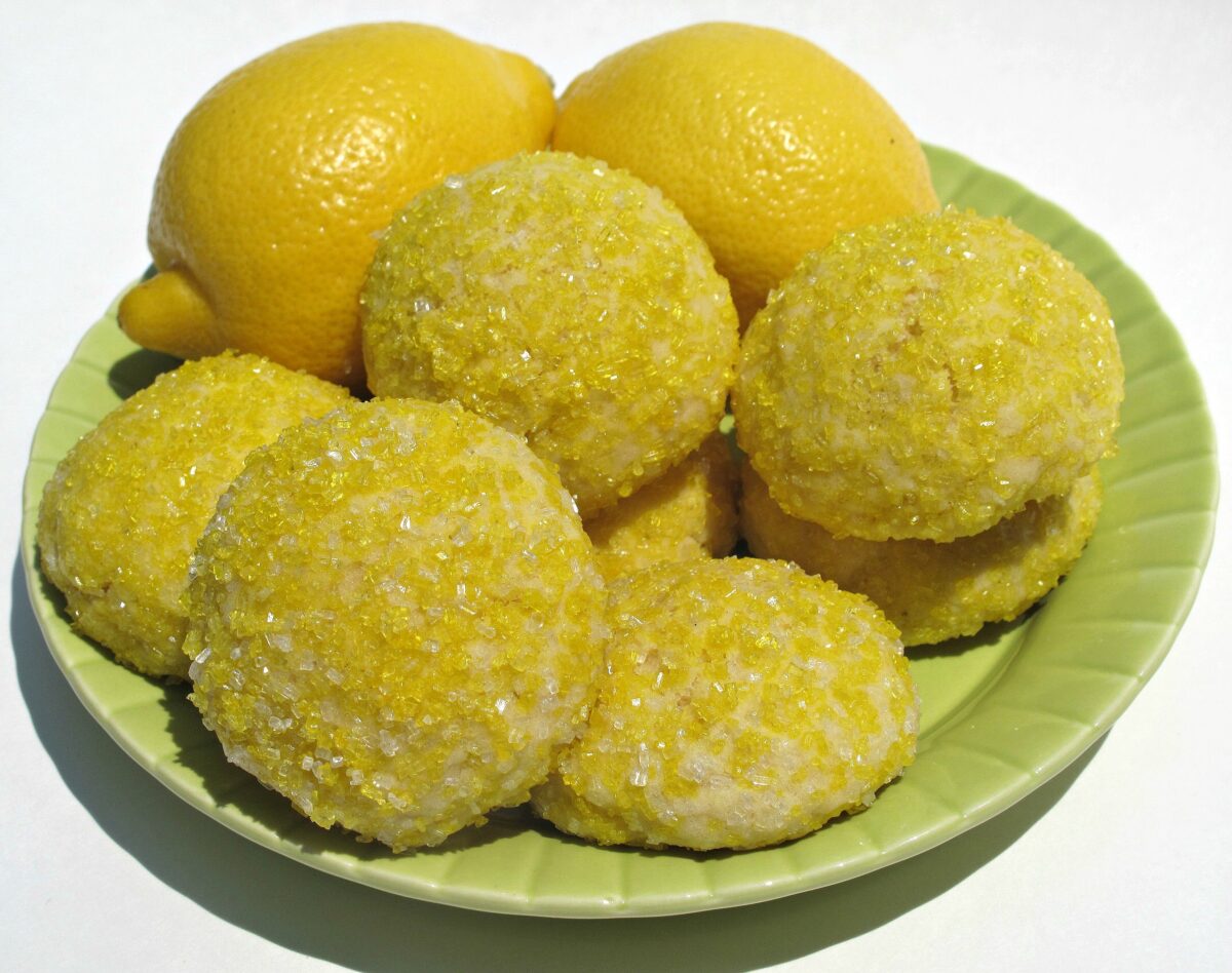 Round lemon pillow cookies covered with yellow sparkling sugar with lemons on a green serving plate.