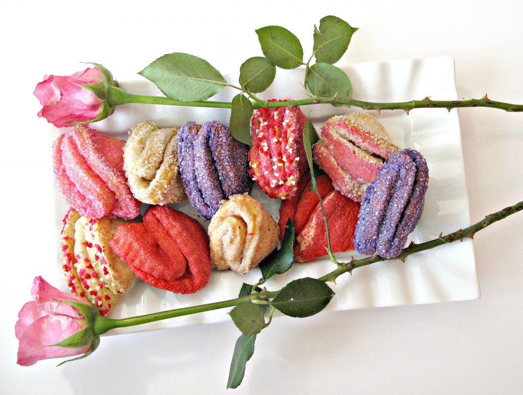 Rose Bud Butter Cookies, folded cookies that look like roses, are coated in pink, red, or purple sparkling sugar arranged on a white platter with two pink roses.