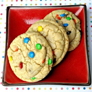 Three M&M cookies on a red plate on top of a polka dotted background.