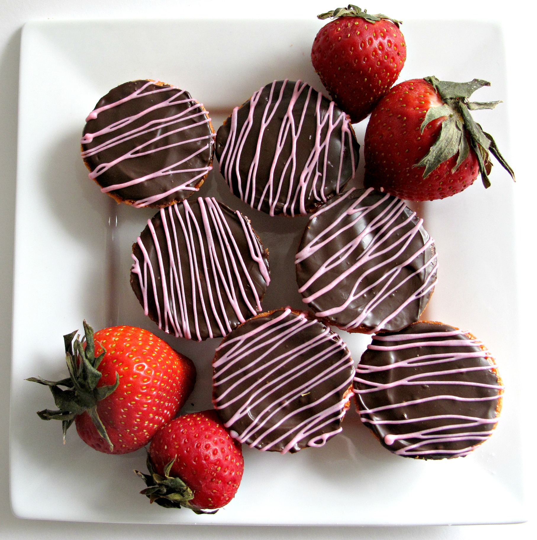 Blondies topped with chocolate and pink zigzags, with 4 strawberries on a square white plate. 