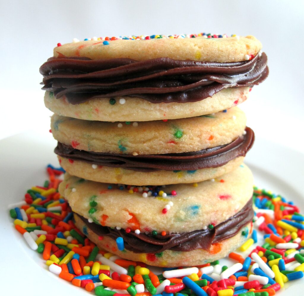 Funfetti Sandwich Cookies in a stack on a plate covered in rainbow colored sprinkles. Each sandwich is two funfetti cookies with chocolate frosting in the middle.