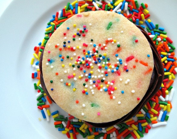 One Funfetti Sandwich Cookie from the top, sitting on a plate covered in sprinkles. The cookie has rainbow colored jimmy sprinkles in the dough and rainbow nonpareils on top.