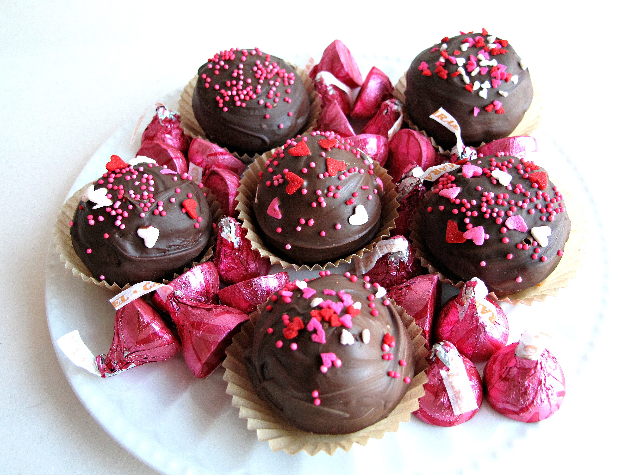 Kiss Cookies, ball-like chocolate cookies dipped in chocolate and sprinkled with Valentine sprinkles, on a plate with pink foil Hershey's kisses
