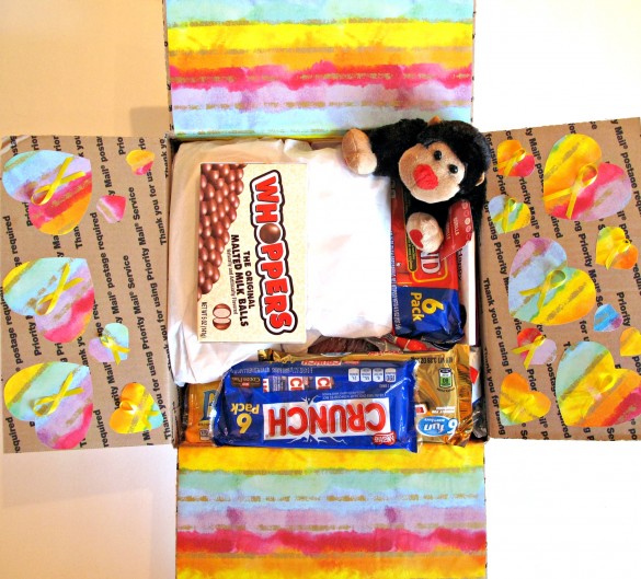 The flaps of the care package are decorated ; 2 are covered with rainbow striped wrapping paper and two have hearts cut from the rainbow paper. Each heart has a yellow "support our troops" ribbon in the middle.