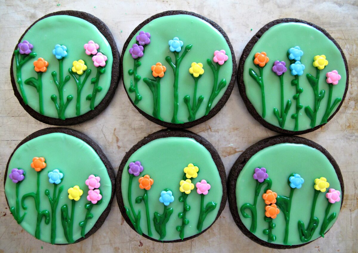 Six chocolate circle cookies decorated with flowers made of icing and sprinkles.