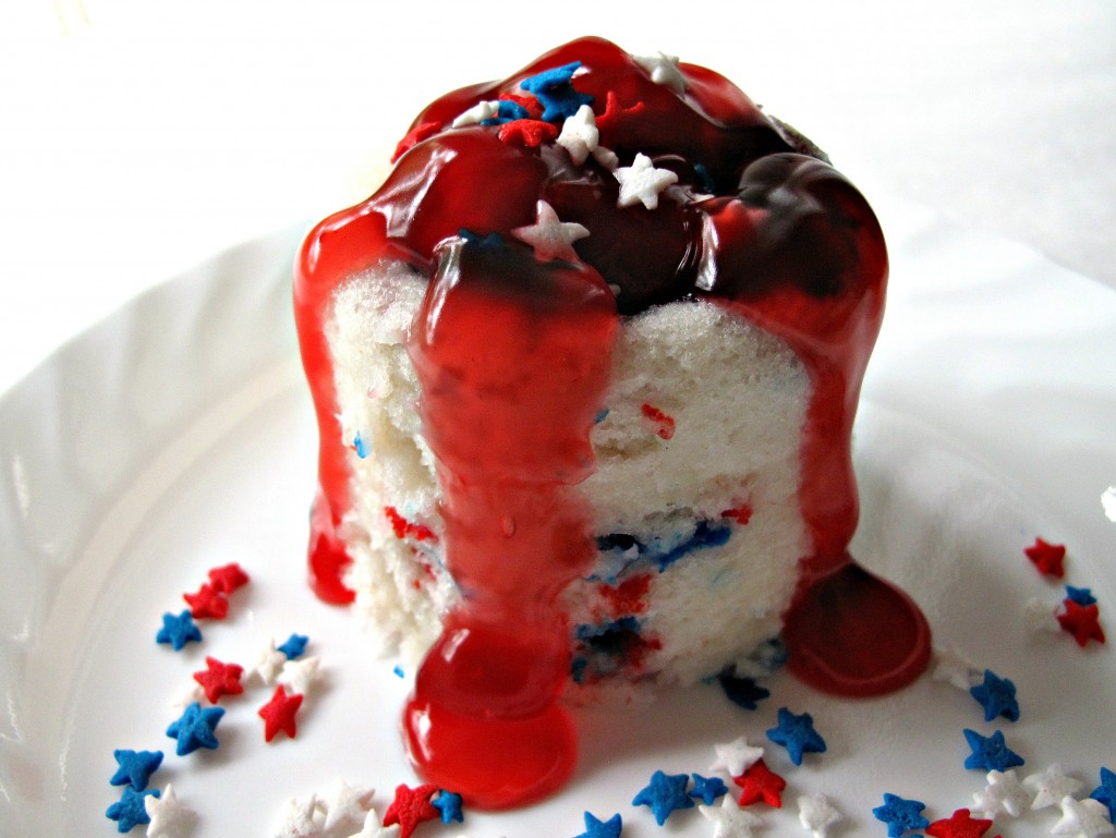 Red, White, and Blueberry Mug Cake, white cake with red and blue funfettin sprinkles topped with strawberry pie filling overflowing down the sides, on a white plate with red, white, and blue star sprinkles.