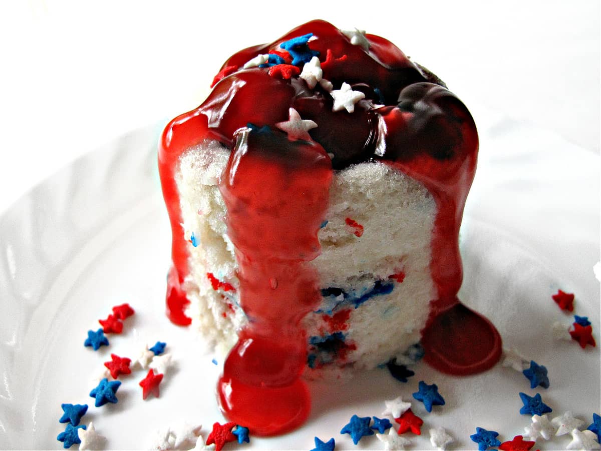 White mug cake with red and blue funfetti sprinkles andstrawberry pie filling overflowing down the sides.