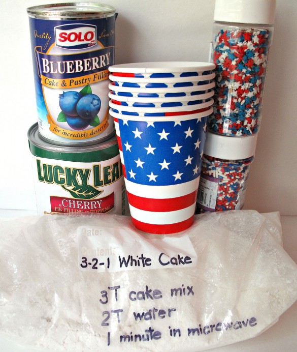 Red, White, and Blueberry Mug Cake ingredients: canned blueberry and cherry pie filling, red/white/blue sprinkles, flag decorated paper cups, large ziplock bag filled with a combination of white cake mix and angel food cake mix. The bag has the directions written on it: 3 tablespoon cake mix, 2 tablespoons water, 1 minute in the microwave.