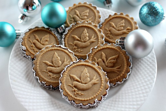 Cookies stamped with a design of a flying dove holding an olive branch, with their bottoms dipped in chocolate and white nonpareil sprinkles, on a round white plate