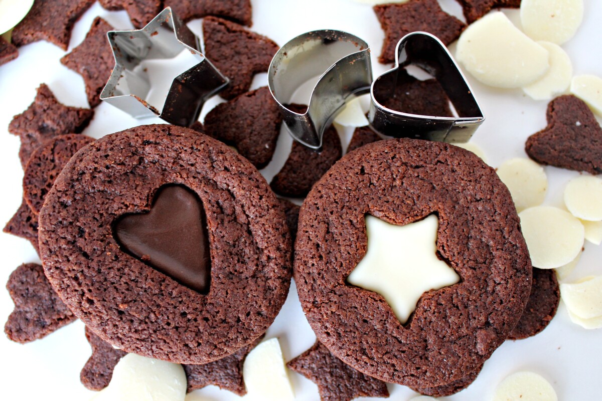 Two sandwich cookies with chocolate filling showing through a heart  and star cutout. 