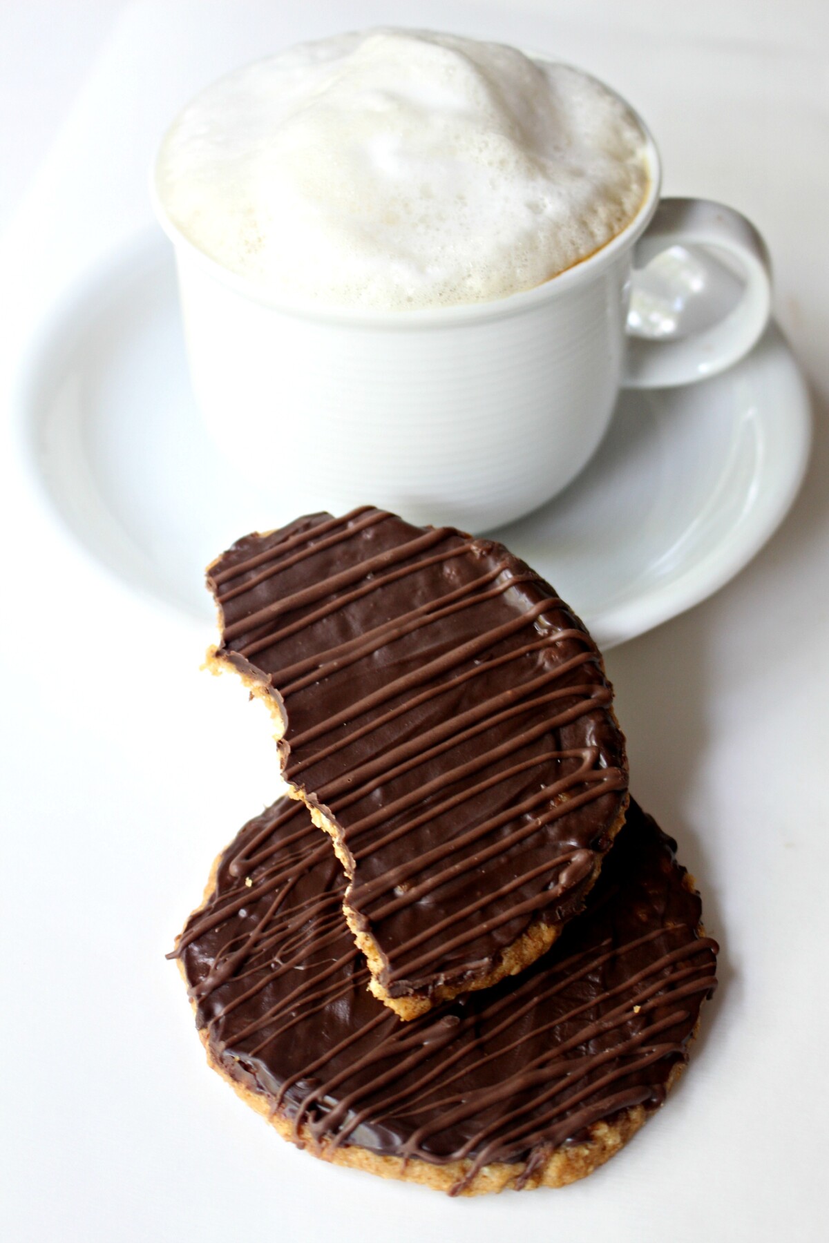 Two cookies in front of a white mug of cappuccino topped with milk foam.