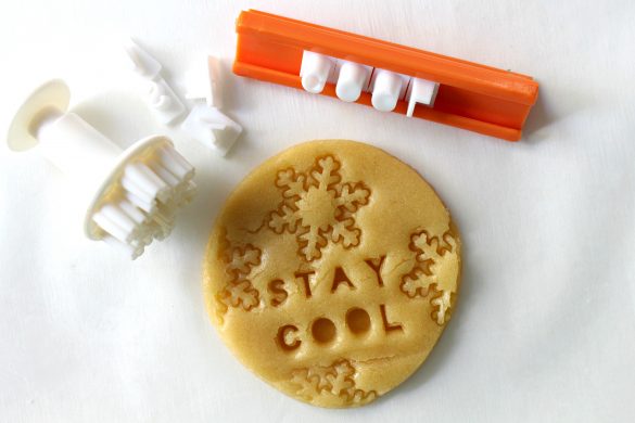 Round cutout sugar cookie dough stamped with snowflakes and the words, "Stay Cool" shown with the snowflake shaped stamp and the word stamp.