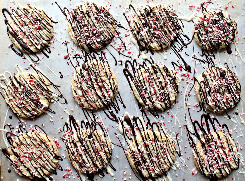 Peppermint Crunch Cookies lined up on wax paper lined baking sheet with drizzles of white and dark chocolate and sprinkled with peppermint candy bits