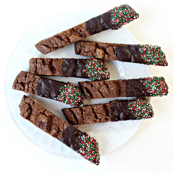Mocha White Chocolate Biscotti on a white plate from above, showing one end of biscotti bare with cracks and the other end dipped in chocolate and nonpareil sprinkles in red, green, and white.