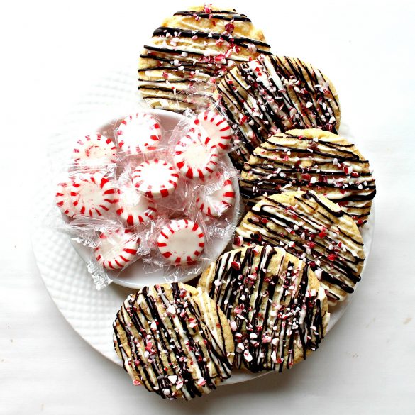 Peppermint Crunch Cookies decorated with drizzle of dark chocolate and peppermint candy bits on a white plate with a bowl of red and white peppermint candies