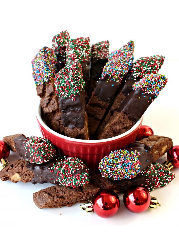 Mocha White Chocolate Biscotti, with one end coated in chocolate and Christmas nonpareil sprinkles, standing up in a red bowl with additional biscotti and red ball Christmas ornaments scattered at the base of the bowl.