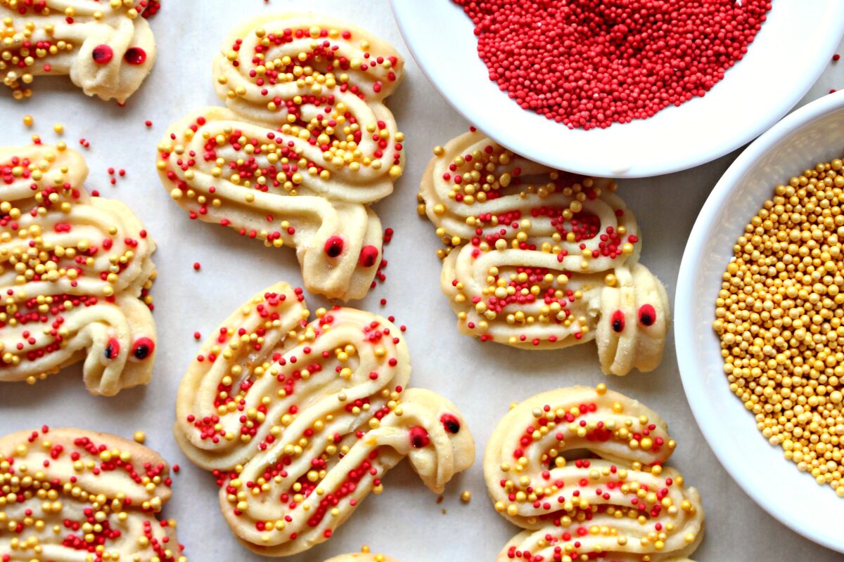 Cookies with red and gold nonpareil sprinkles on a white surface with two bowls of sprinkles.