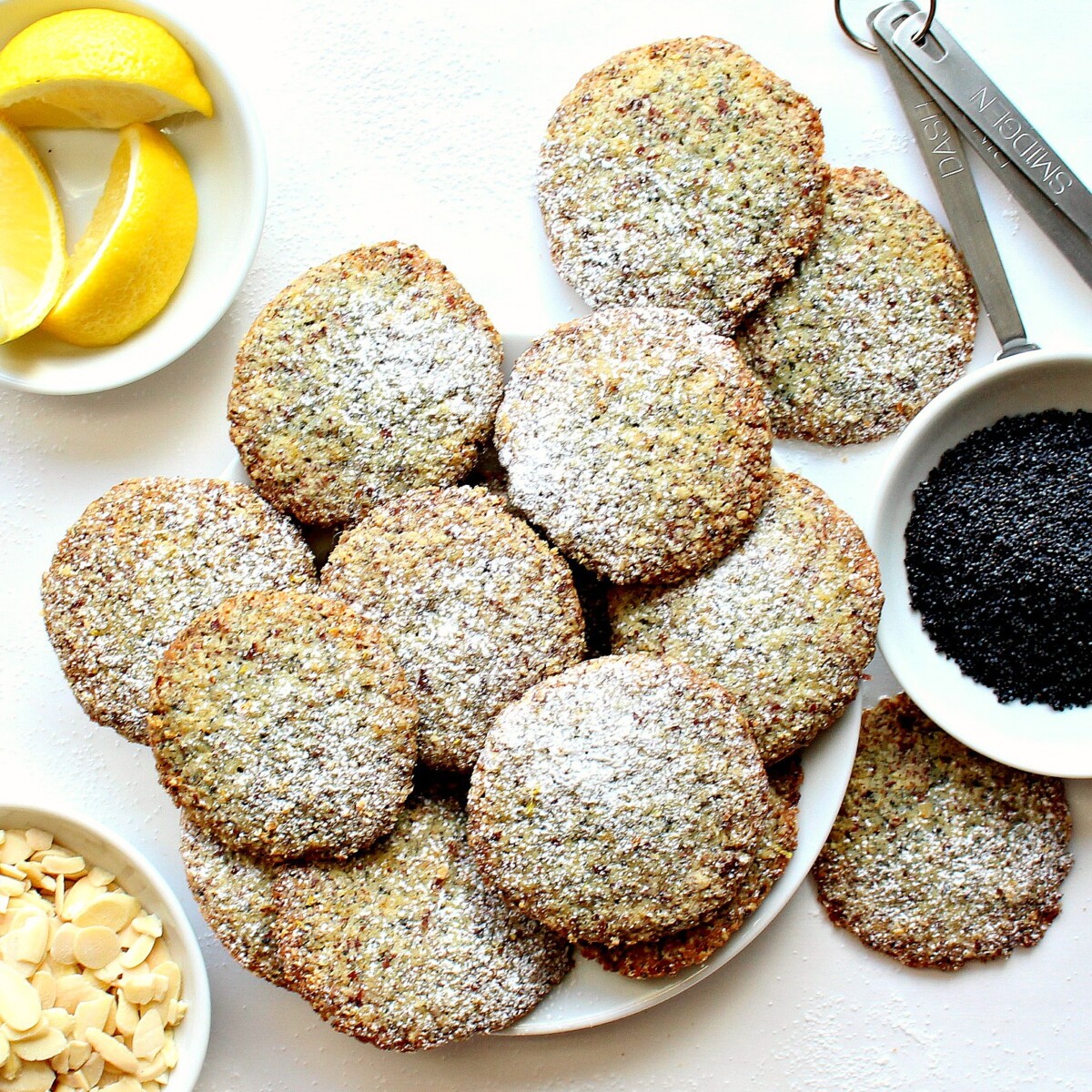  Cookies on a serving plate with bowls of poppy seeds, slivered almonds, and lemon slices.