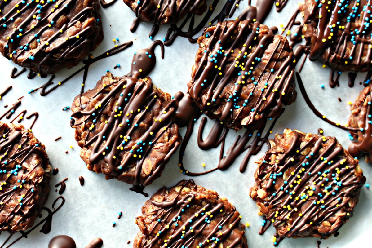No-Bake Chocolate Peanut Butter Banana Cookies on wax paper immediately after applying chocolate drizzle and nonpareil sprinkles. Drizzle extends beyond the cookies and onto the paper.