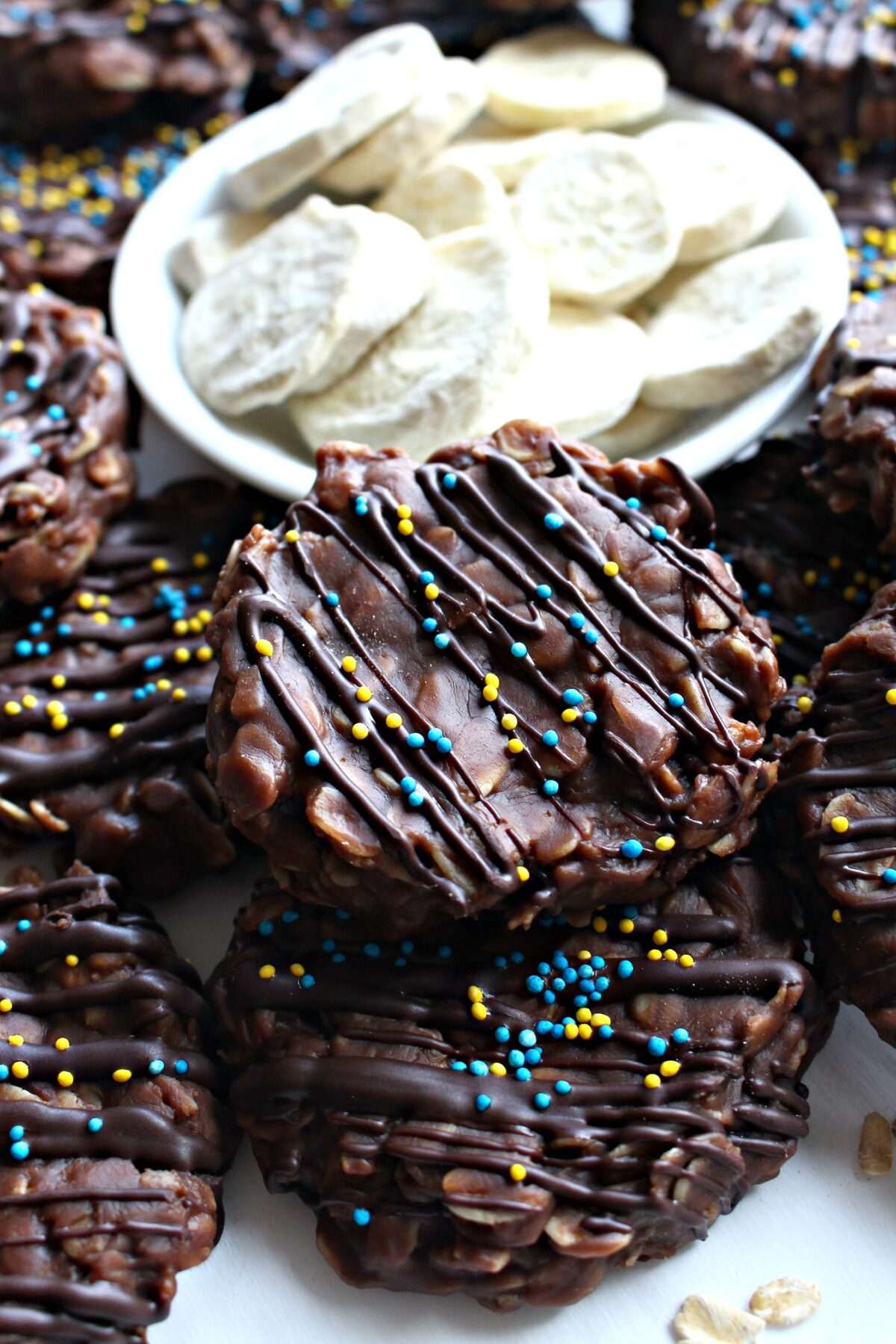 No-Bake Chocolate Peanut Butter Banana Cookies, thick discs of chocolate and oats, drizzled with chocolate and sprinkled with yellow and blue nonpareils.