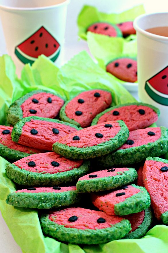 Watermelon Sugar Cookies in a serving dish lined with bright green tissue paper. The cookies are half circles that look just like slices of watermelon. They are bright pink with a green "rind" edge and three black "seeds" all made of colored cookies dough.