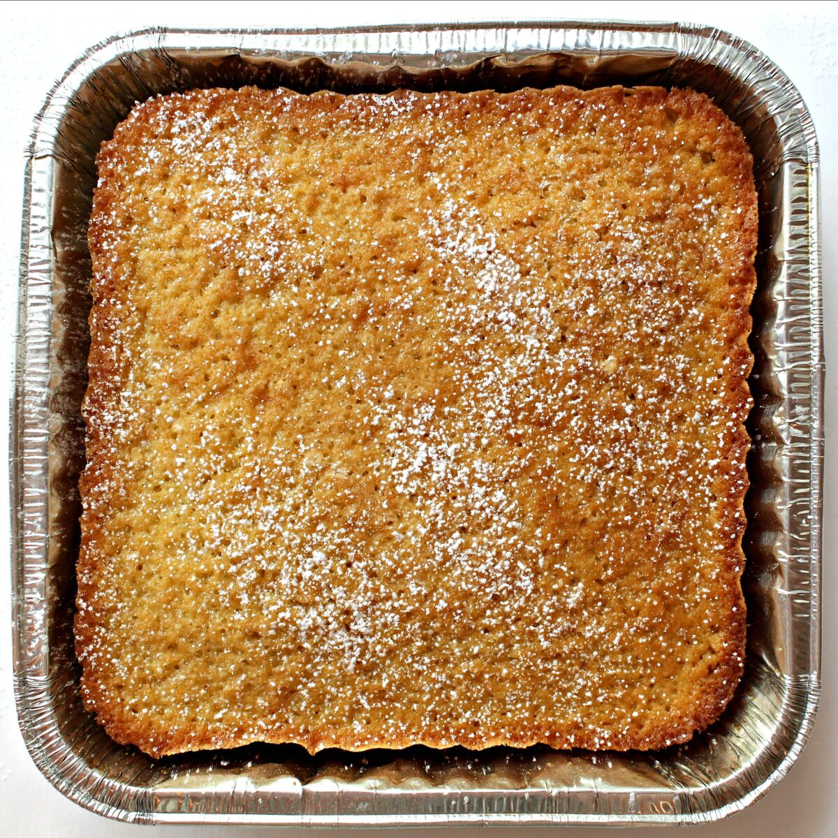 Coconut Milk Snack Cake in a foil pan with powdered sugar sprinkled on top.