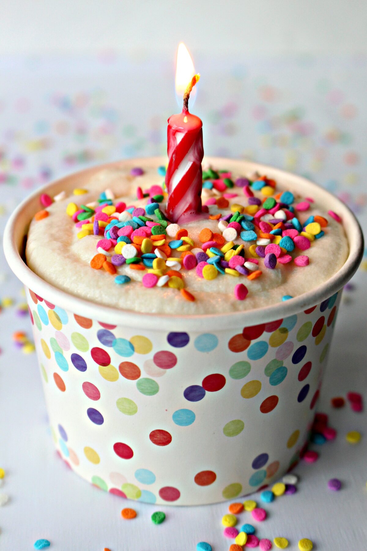 Individual frosted microwave cake in a paper cup with confetti sprinkles and a lit candle.