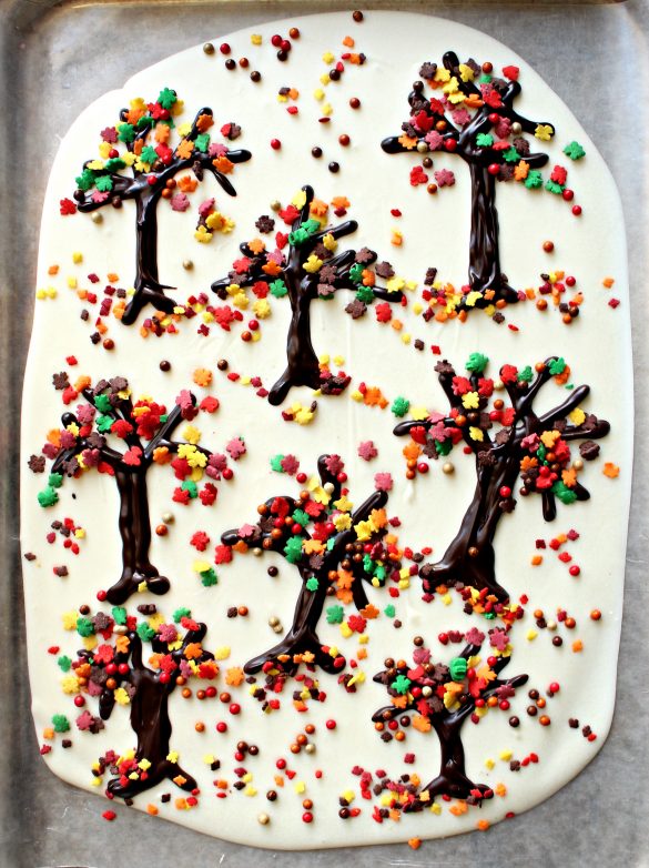 Thanksgiving White Chocolate Bark is melted white chocolate spread into a large rectangle then melted dark chocolate is piped on top as trees scattered around the rectangle. Leaf sprinkles in green, yellow, red, orange, and brown are sprinkled onto the trees to look like fall leaves.