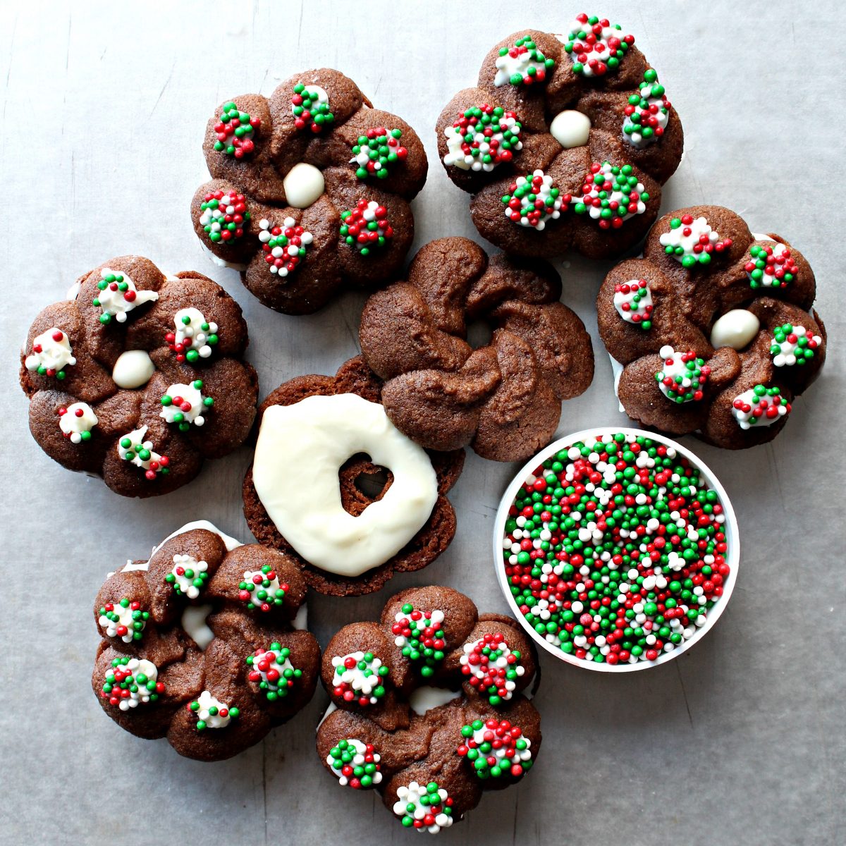 Chocolate Spritz Cookies in wreath shapes, sandwiched with white chocolate, topped with red and green nonpareils.