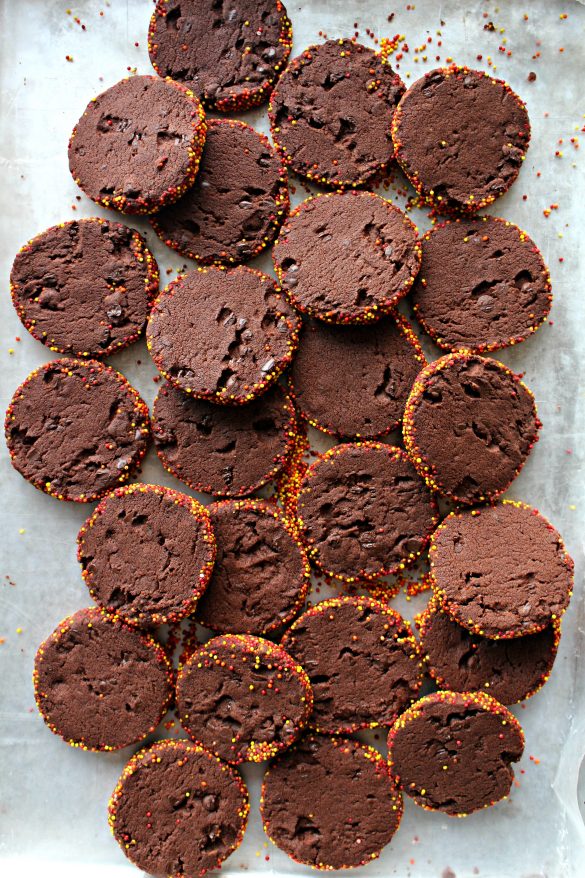 Spiced Chocolate Shortbread Cookies are sliced from a dough log that is covered in orange and yellow nonpareil sprinkles.