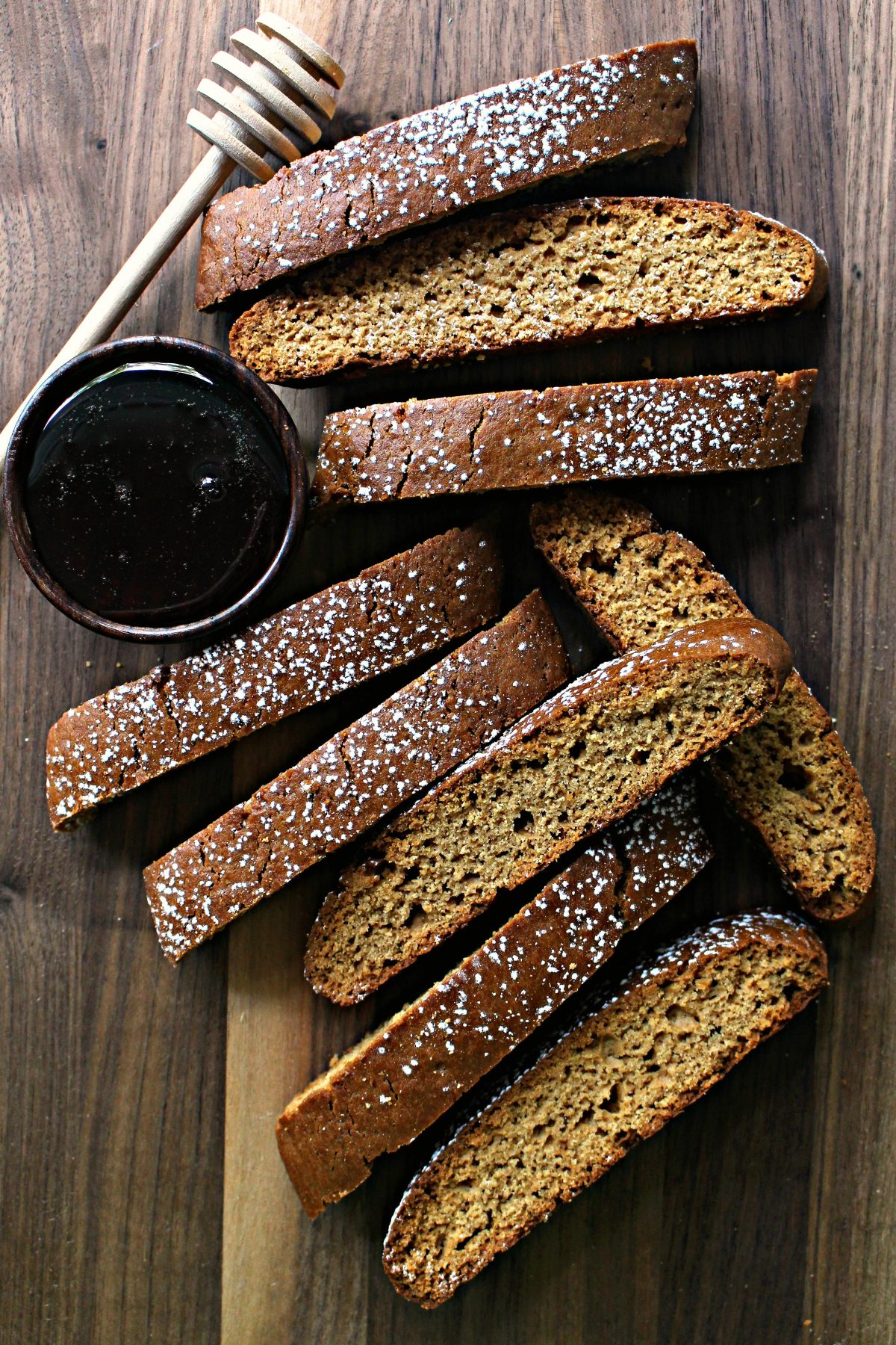 Biscotti with powdered sugar on cutting board with a bowl of honey and a honey server.