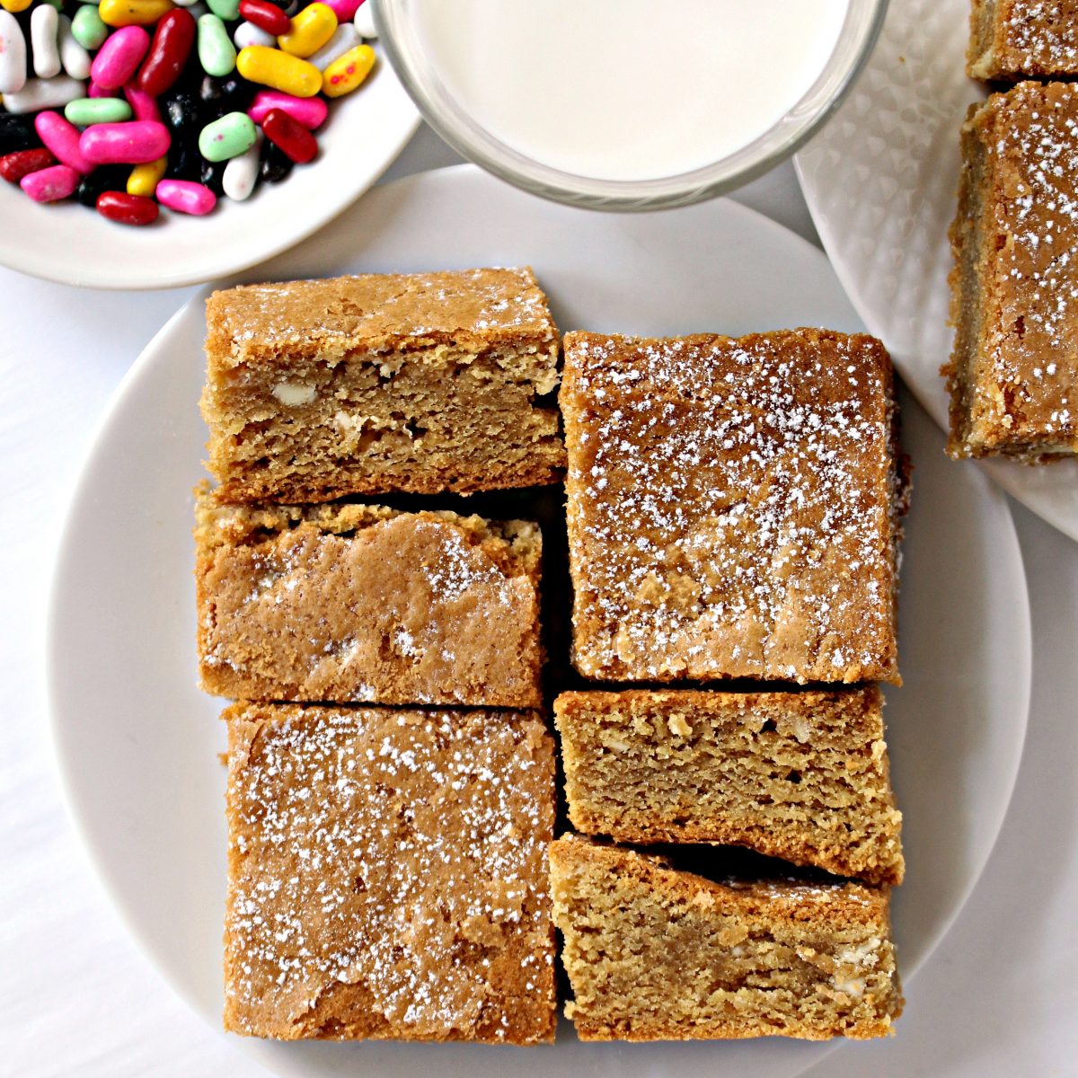 Cookie bars on a plate with a glass of milk and a bowl of licorice candy.