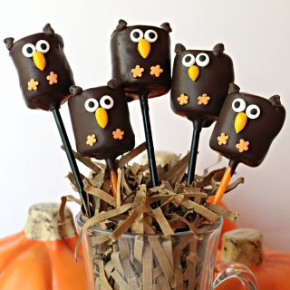 Five owl marshmallow pops on sticks in a cup filled with shredded paper like a nest