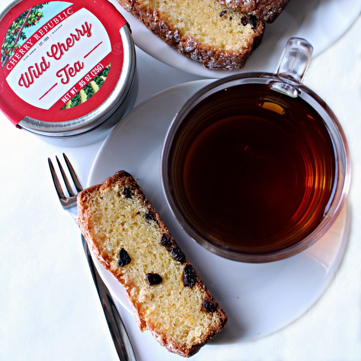 Cup of tea on a saucer with a slice of cake and a tin of cherry tea.