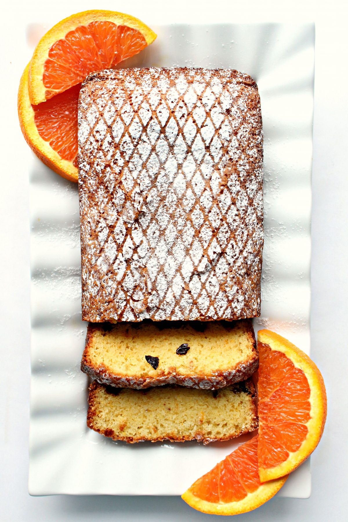Orange loaf cake topped with powdered sugar grid design on a white platter with orange slices.