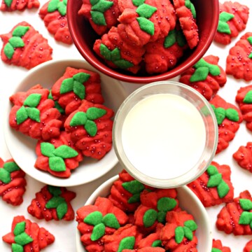 Red strawberry shaped cookies with green leaves on plates and a bowl with a glass of milk