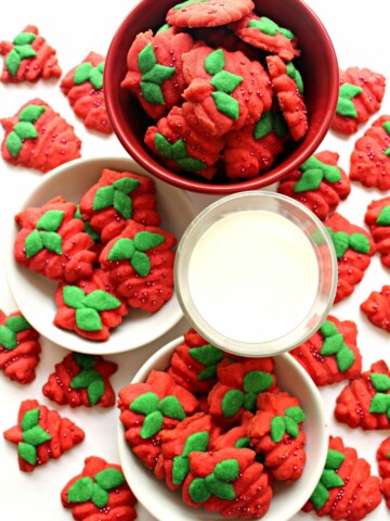 Red strawberry shaped cookies with green leaves on plates and a bowl with a glass of milk