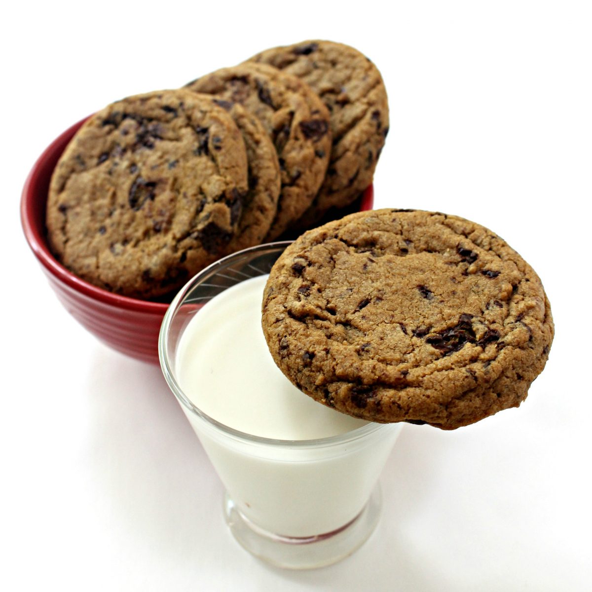 Cookie balanced on rim of glass of milk with a bowl of cookies in the background