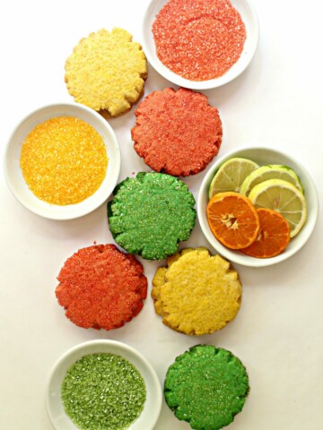 sugar cookies on white background with white bowls of colored sugar and sliced citrus fruit