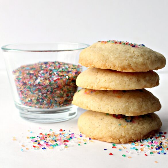 Stack of cookies on white surface with a  glass of multicolored decorating sugar in the background.