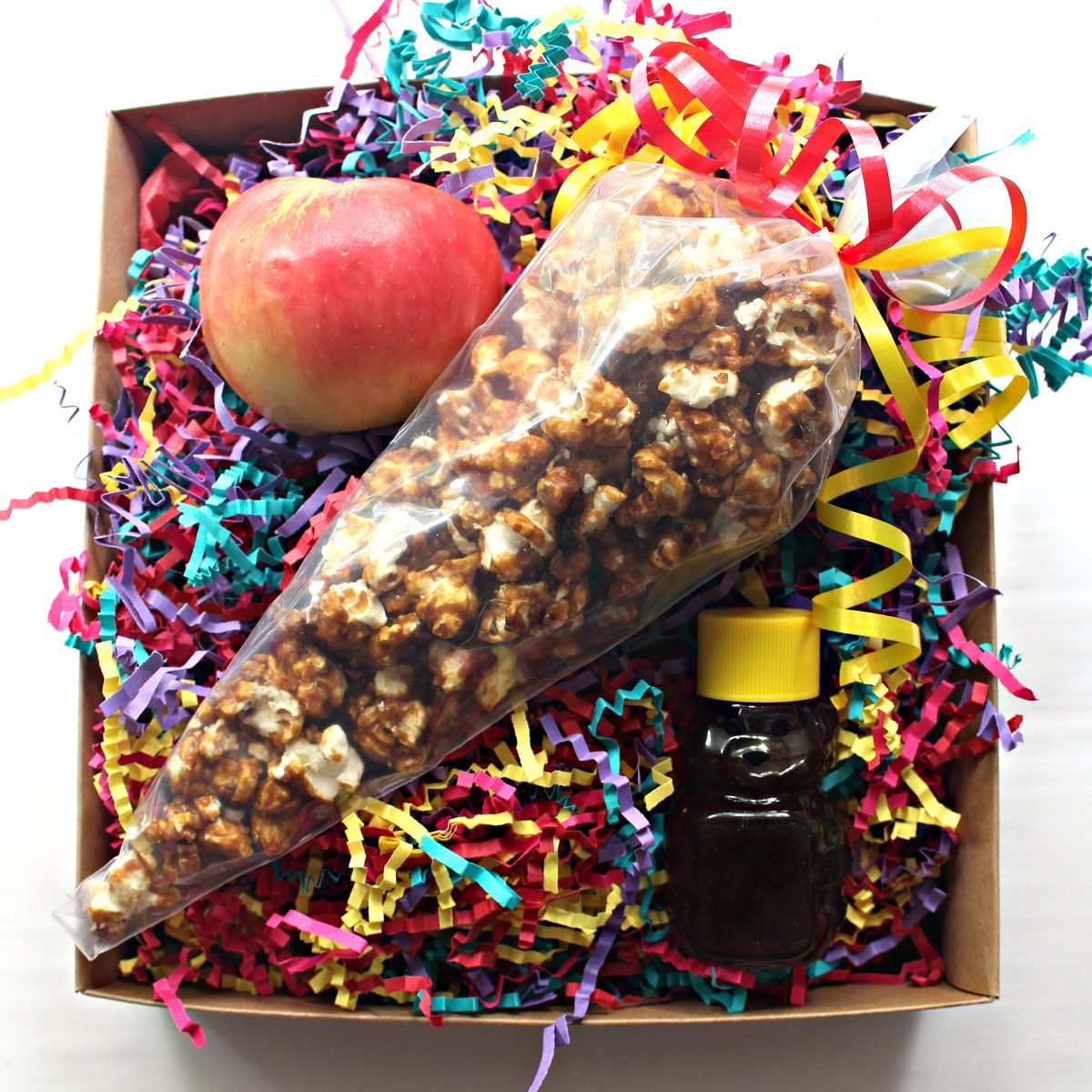 Gift package with rainbow raffia, apple, honey bear, and a clear cone bag filled with popcorn.