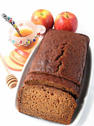 Sliced loaf of Honey Cake on a platter next to a honey jar and apples.