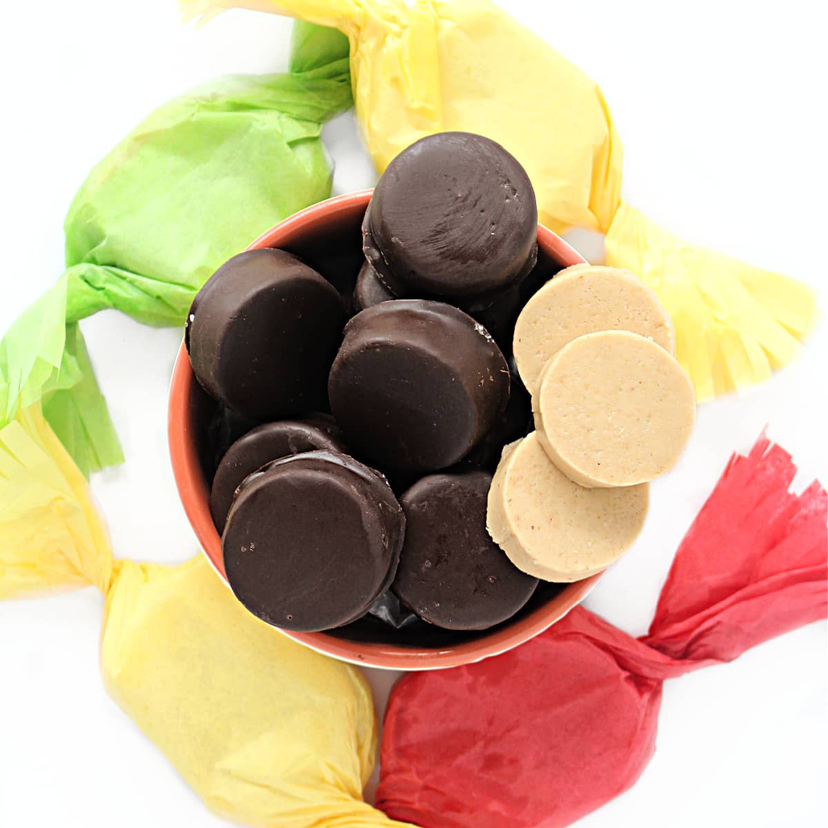 Bite sized, chocolate coated mazapan candy discs in a bowl surrounded by tissue paper wrapped candies.