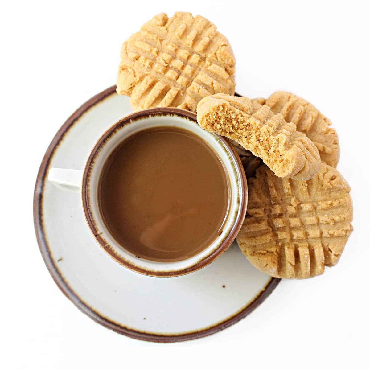 Coffee cup with peanut butter cookies on the saucer with one cookie showing interior crumb.