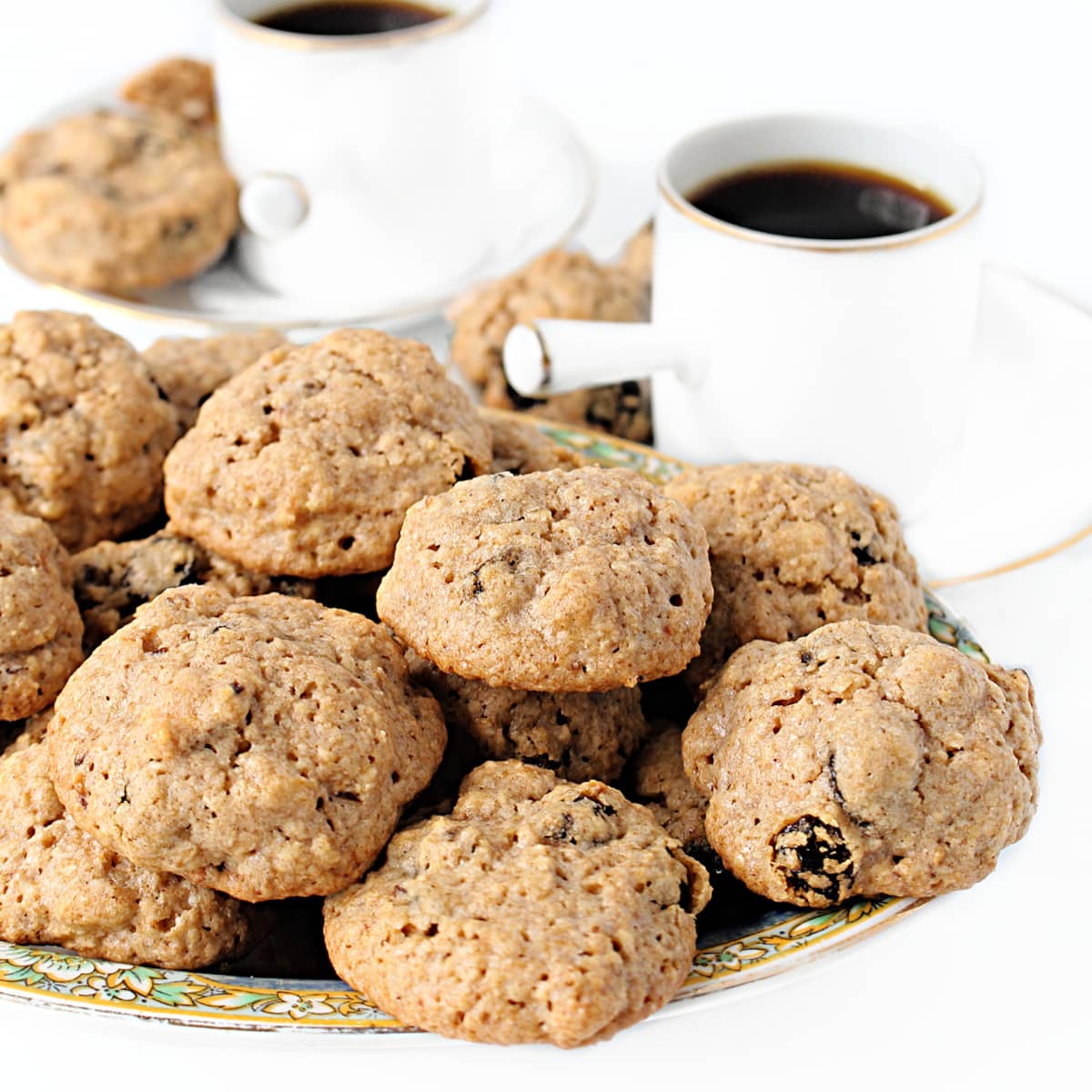 Closeup of small, mounded mock oatmeal cookies on a platter with coffee cups in the background.