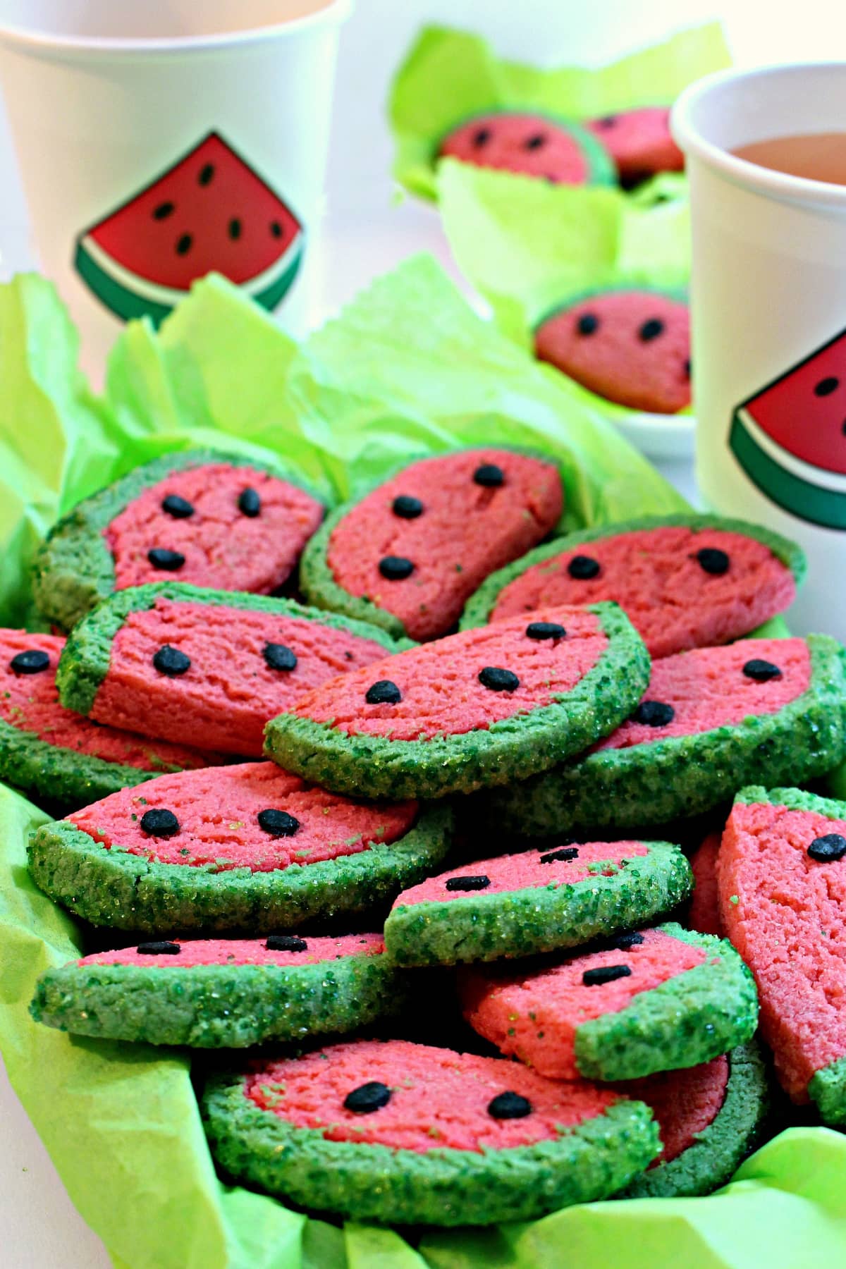 Platter of sugar cookies resembling watermelon slices andwatermelon slice decorated paper cups in the background.