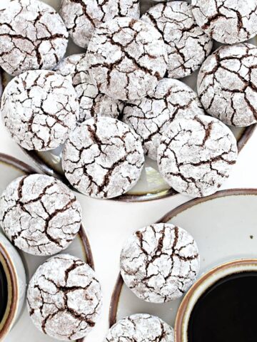 Plate of chocolate crinkle cookies covered in powdered sugar with cracks showing chocolate cookie inside.