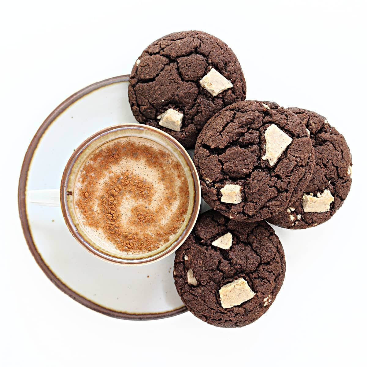 Fudgy Cappuccino Cookies with crackled tops and chunks of cappuccino white chocolate on a saucer next to a mug of coffee.